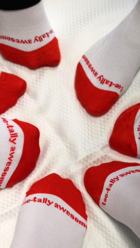 A bunch of feet wearing white socks with a red toe. Along the sock it reads Toe-tally Awesome